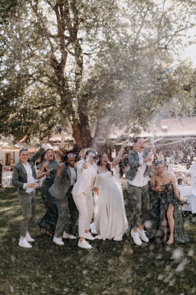 Eine Hochzeitsgesellschaft feiert im Freien, lässt Champagner knallen und lacht inmitten von Seifenblasen und Sonnenlicht.