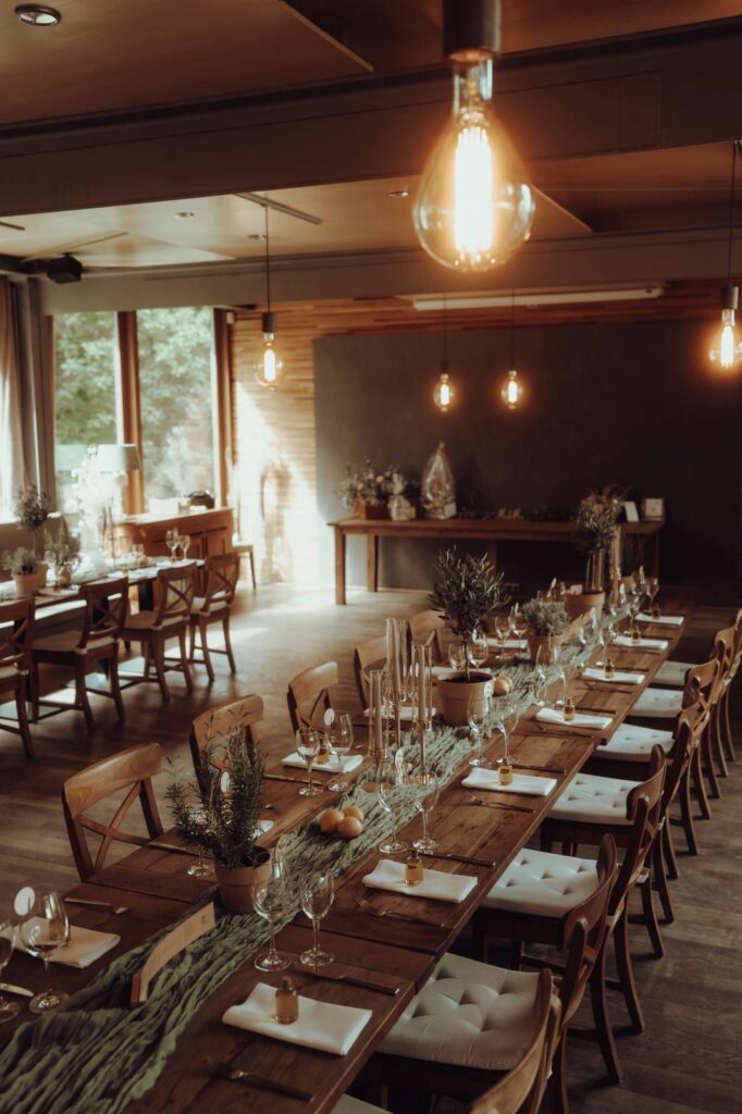 Elegantes Esszimmer mit rustikalen Holztischen, die für eine Mahlzeit gedeckt sind, dekoriert mit Kerzen und Grünzeug.