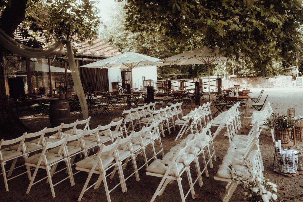 Weiße Stuhlreihen im Freien unter Bäumen, aufgestellt für eine Hochzeit oder eine Veranstaltung mit Sonnenschirmen als Schattenspender.