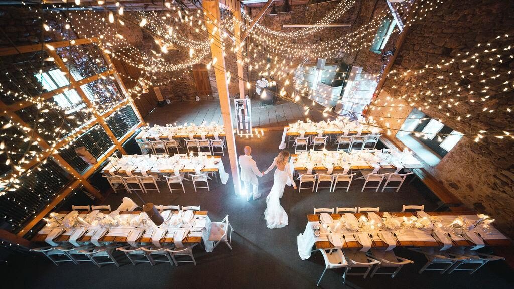 Ein Paar geht Hand in Hand durch eine warm beleuchtete, mit Lichterketten und Banketttischen geschmückte Hochzeitslocation.