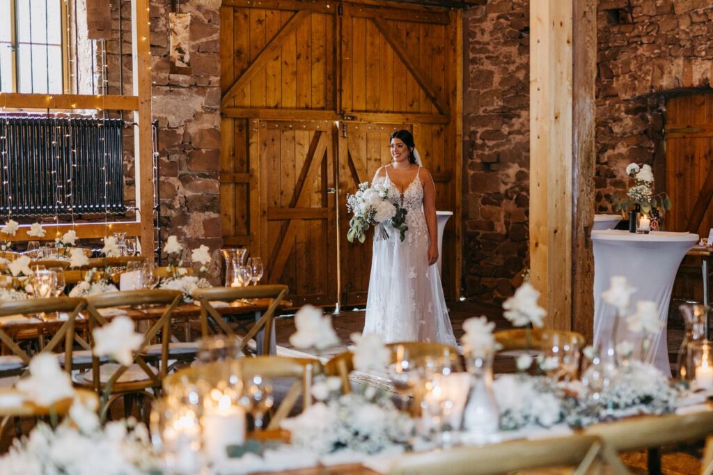 Eine Braut in einem weißen Kleid steht in einer rustikalen, mit Blumen und Kerzen geschmückten Scheune.