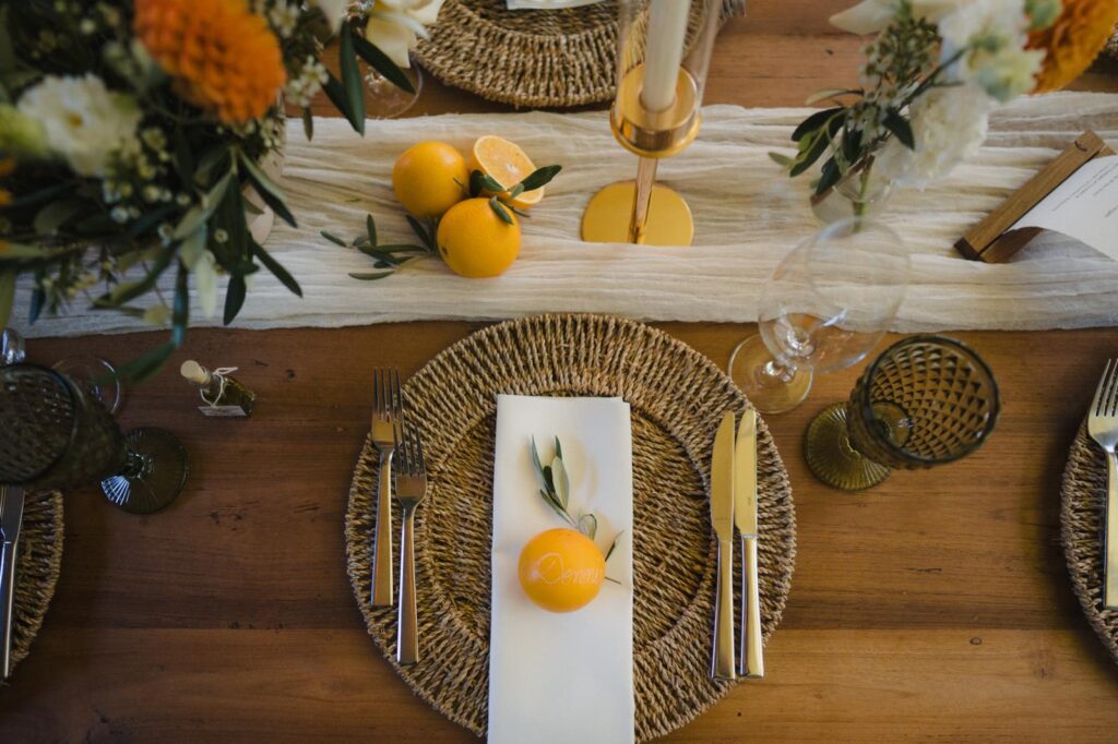Gedeckter Tisch mit gewebtem Tischset, Besteck, Serviette, orangefarbenen Früchten und Blumendekor auf einer Holzfläche.