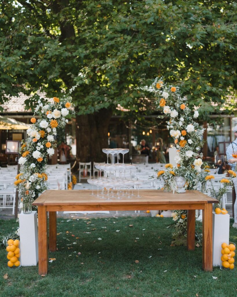 Ein Holztisch mit einem Turm aus Champagnerglas, umgeben von orangefarbenen und weißen Blumenbögen im Freien - Hochzeitslocation mit Wedding Planner Mia Weydert