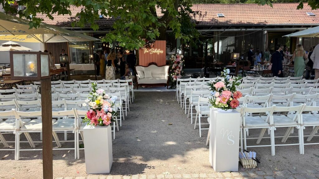 Hochzeit im Freien mit weißen Stühlen, Blumenarrangements und einer dekorierten Kulisse mit einem Sofa.