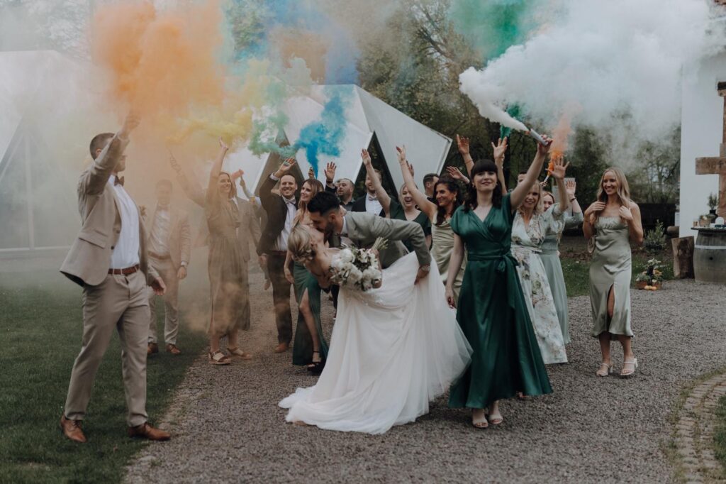 Braut und Bräutigam küssen sich, während ihre Freunde bei einer Hochzeit im Freien mit bunten Rauchbomben jubeln.