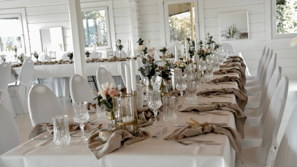 Elegante Banketttische mit Gläsern, beigen Servietten und blumigen Tafelaufsätzen in einem hellen, weißen Raum.
