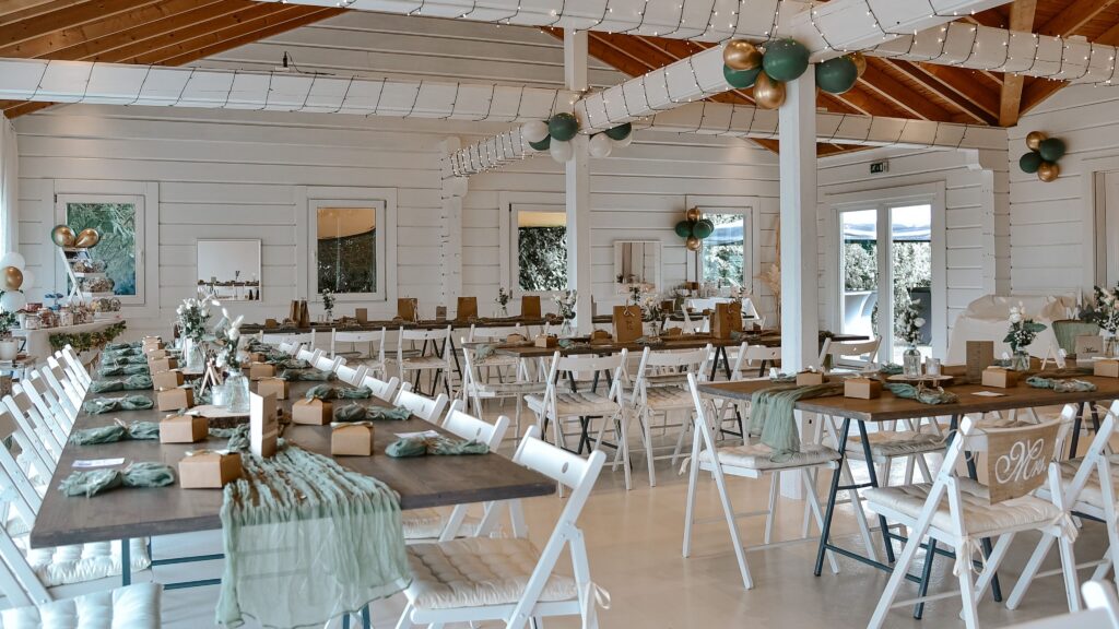 Dekorierter Bankettsaal mit Holzbalken, langen Tischen, weißen Stühlen, grünen Akzenten und Lichterketten.