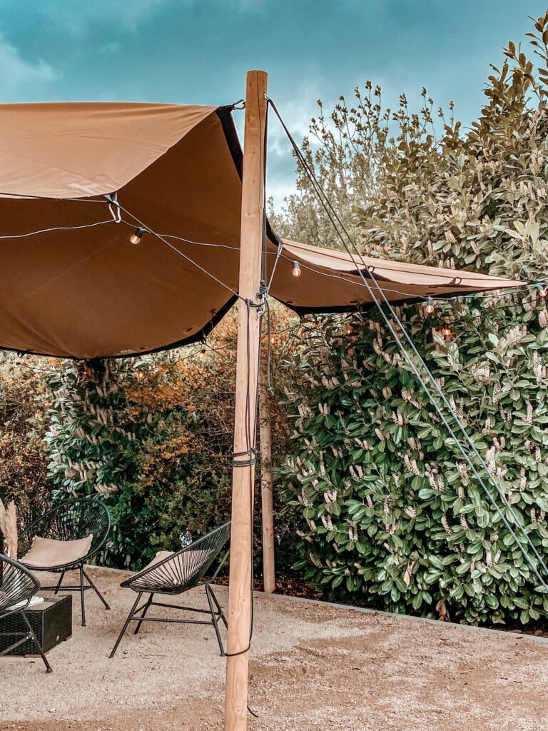 Sitzbereich im Freien mit Stühlen unter einem braunen Vordach, umgeben von üppigen grünen Büschen.