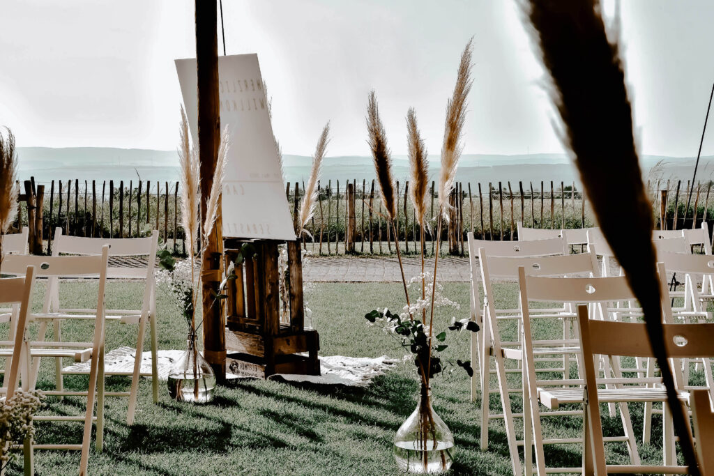 Hochzeit im Freien mit weißen Stühlen, Pampasgras in Vasen und einer hölzernen Staffelei auf einem grasbewachsenen Rasen.