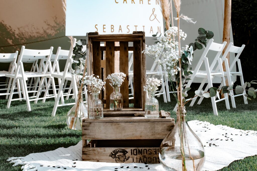 Holzkisten mit Glasvasen und weißen Blumen auf einem Teppich, mit weißen Stühlen, die für eine Hochzeit im Freien aufgestellt sind.