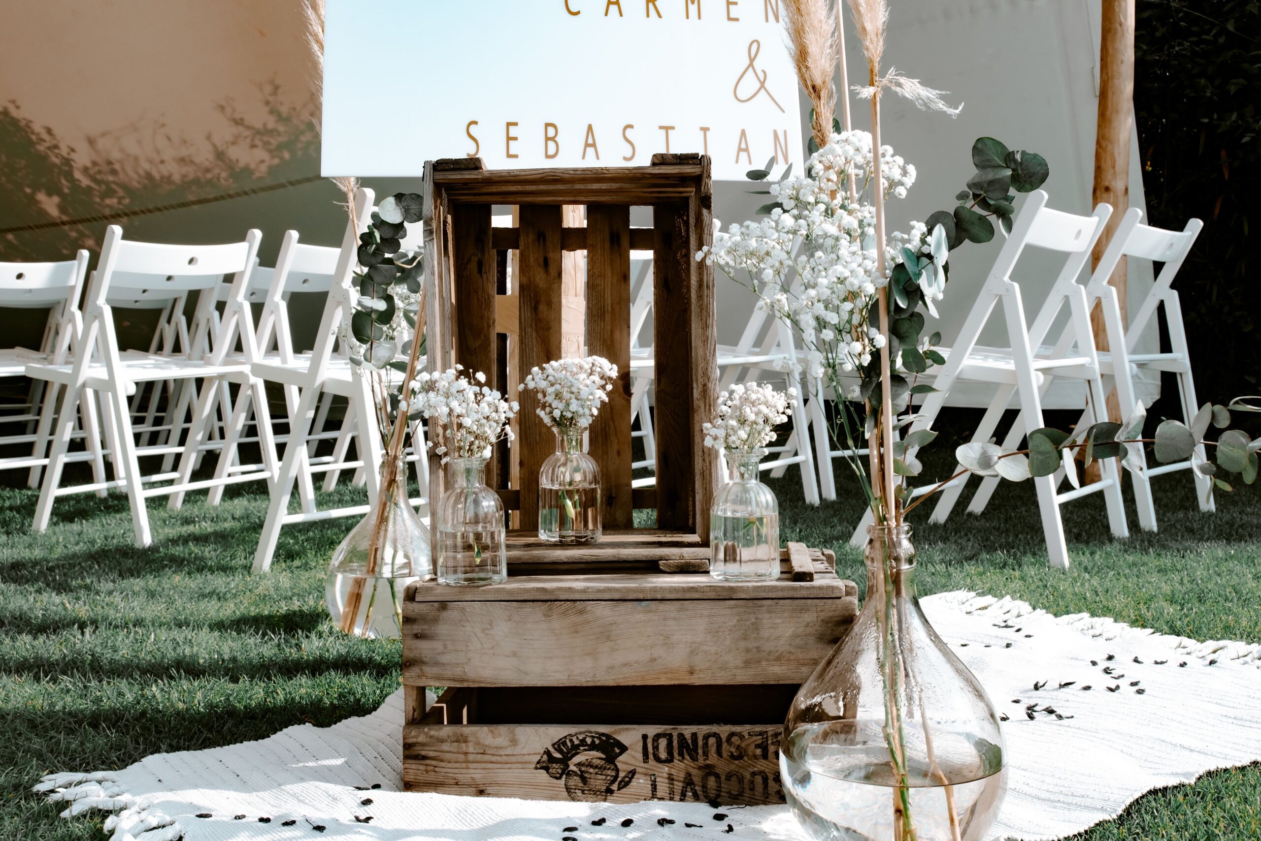 Holzkisten mit Glasvasen und weißen Blumen auf einem Teppich, mit weißen Stühlen, die für eine Hochzeit im Freien aufgestellt sind.
