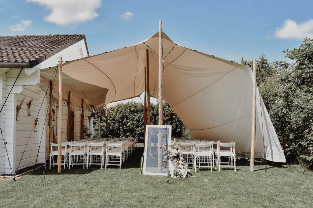 Hochzeitsempfang im Freien unter einem weißen Zelt mit weißen Stühlen und einem geblümten Willkommensschild.