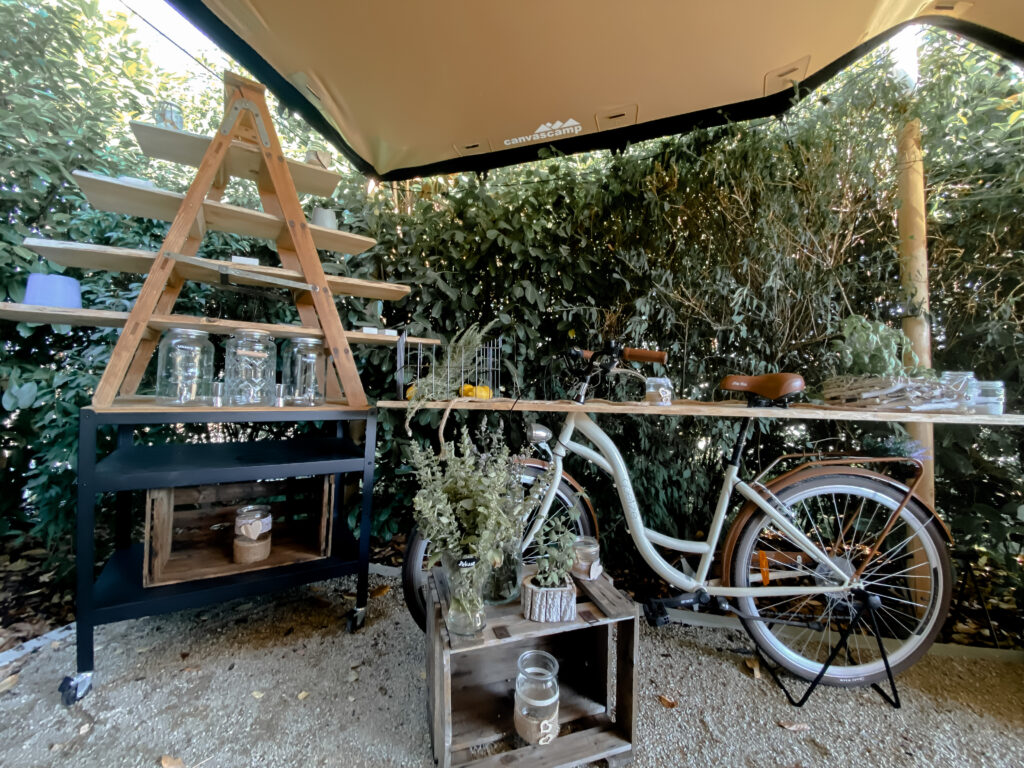 Einrichtung im Freien mit einem Fahrrad, Holzregalen, Glasgefäßen und Grünzeug vor einem grünen Hintergrund.