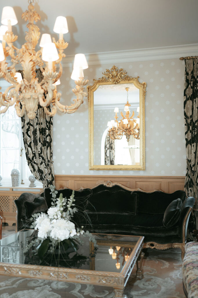 Elegantes Wohnzimmer mit einem goldenen Kronleuchter, einem verzierten Spiegel, einem schwarzen Samtsofa und einem Blumenarrangement auf einem Glastisch.