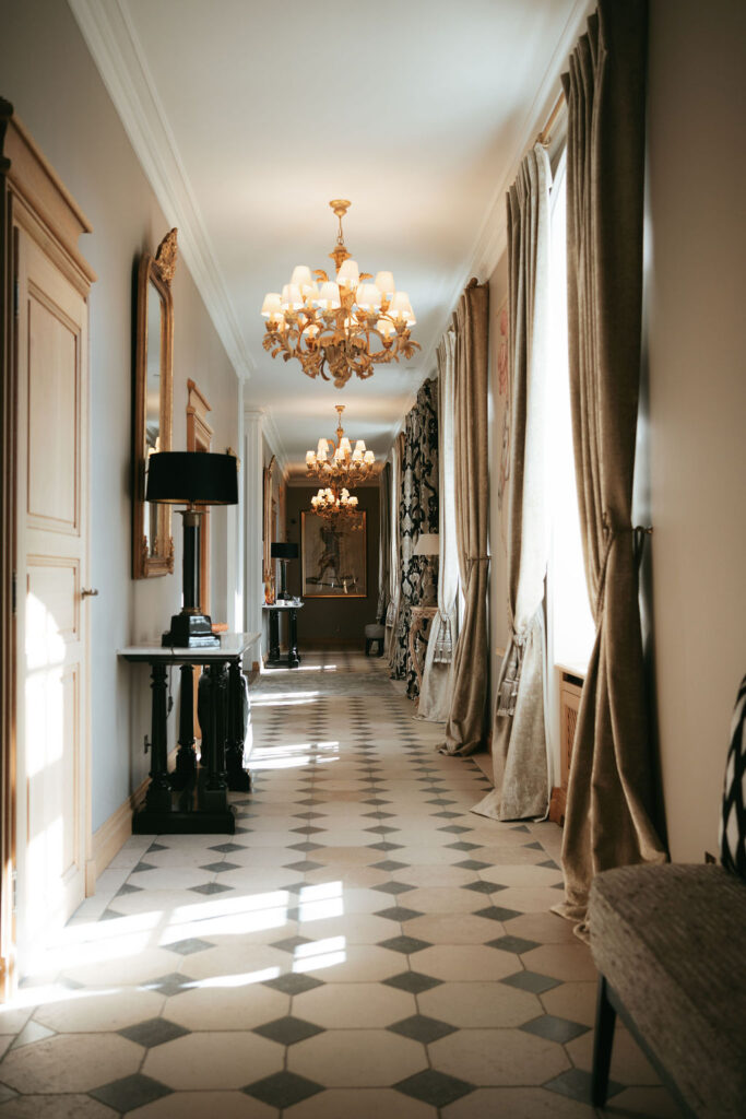 Eleganter Flur mit Kronleuchtern, hohen Fenstern mit Vorhängen, Spiegeln und Fliesenböden, beleuchtet von natürlichem Sonnenlicht.