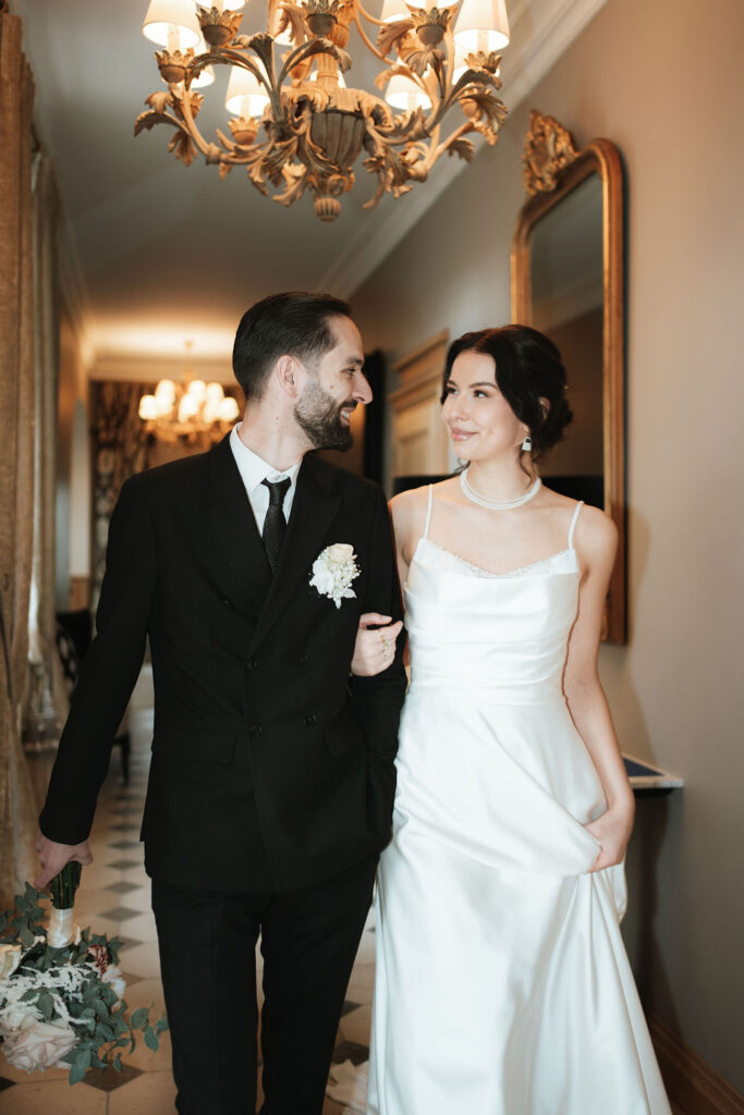 Eine Braut und ein Bräutigam lächeln sich an, während sie Arm in Arm durch einen eleganten Flur gehen.