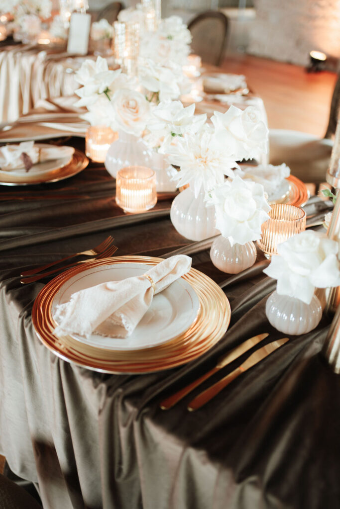 Elegant gedeckter Tisch mit goldumrandeten Tellern, weißen Servietten und weißen Blumen in Vasen auf einem dunklen Tischtuch.